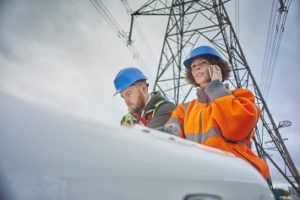 male and female engineers beneath an electricity pylon looking at plans on the bonnet of their van . The female engineer is on the phone.